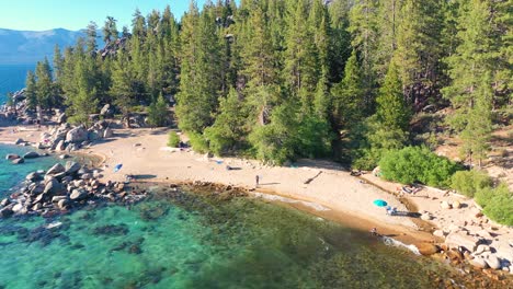 Public-Beach-At-Lake-Tahoe,-California-with-Thick-Pine-Tree-Woods-in-the-Background,-People-Enjoying-Sun,-Clear-Blue-Waters,-and-Large-Rock-Boulders---aerial-drone-pullback