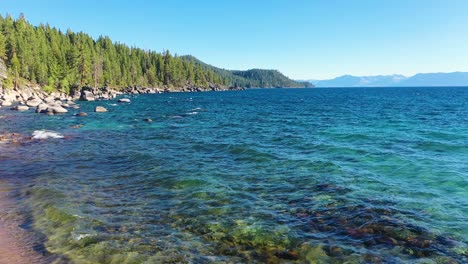 Crystal-Clear-Water-on-Shoreline-Of-Lake-Tahoe-with-Waves-Peacefully-Rippling-on-Beach-In-The-Sierra-Nevada-Mountains-with-Rock-Boulders-and-Pine-Tree-Forest---wide-shot