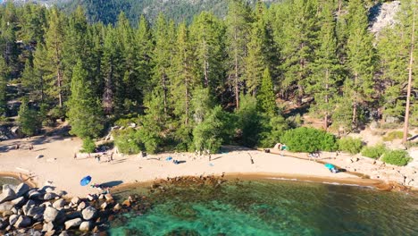 Aerial-Drone-View-of-Public-Beach-At-Lake-Tahoe,-California-with-People-Walking,-Thick-Pine-Tree-Forest-in-Background,-Clear-Blue-Waters,-and-Large-Rock-Boulders