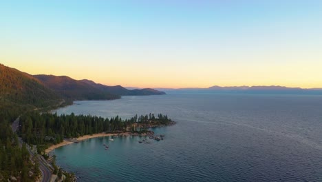 Aerial-Drone-View-of-a-Beautiful-Sunrise-Over-Beach-at-Lake-Taho-with-a-Road-or-Highway-Going-Through-a-Pine-Tree-Forest-with-Clear-Blue-Water-and-Mountains-in-the-Background
