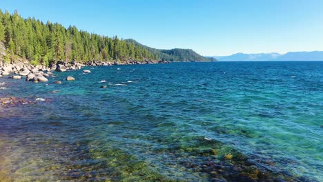 Ruhiges-Blaues-Wasser-Am-Ufer-Des-Lake-Tahoe-Mit-Wellen,-Die-Friedlich-Am-Strand-In-Den-Sierra-Nevada-bergen-Mit-Felsbrocken-Und-Kiefernwald-Plätschern---Breite-Aufnahme