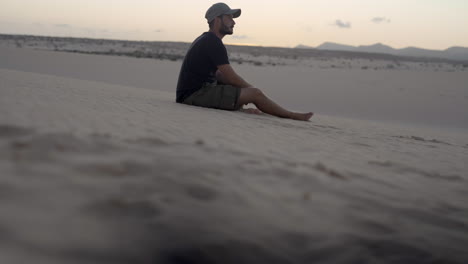 Junger-Abenteurer-Barfuß-Reisender-Sitzt-Allein-In-Sandwüstendünen-Während-Des-Sonnenuntergangs,-Trockene-Malerische-Landschaft-In-Abgelegenen-Bergen-Fuerteventura-Kanarische-Insel-Spanien