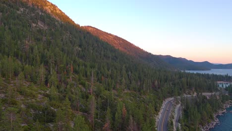 Cars-Driving-on-Mountain-Highway-Road-Through-Jeffrey-Pine-Tree-Forest-and-Blue-Waters-of-Lake-Tahoe-At-Sunset-In-California---aerial-drone-shot