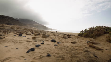 Isla-Fuerteventura,-España-Canaria,-Caminando-Rápido-En-La-Playa-De-Arena-Con-Una-Mujer-Solitaria-Parada-Sola-A-Distancia-Cerca-Del-Agua-Del-Océano