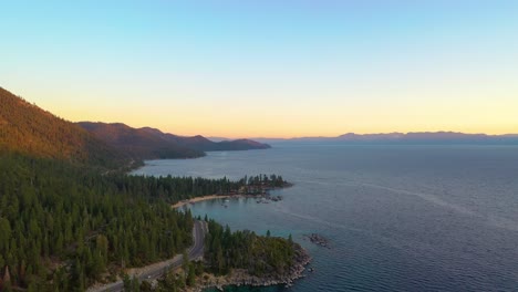 Aerial-Drone-View-of-Beautiful-Lake-Taho-Water-and-Forest-at-Sunrise-with-a-Road-or-Highway-Going-Through-a-Pine-Trees-and-Mountains-in-the-Background-over-a-Beach