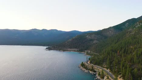 Vista-Aérea-De-Drones-De-Autos-Conduciendo-En-La-Carretera-Junto-Al-Lago-Con-Montañas-Y-Agua-Azul-Del-Lago-Tahoe-En-La-Sierra-Nevada-En-Un-Día-Nublado