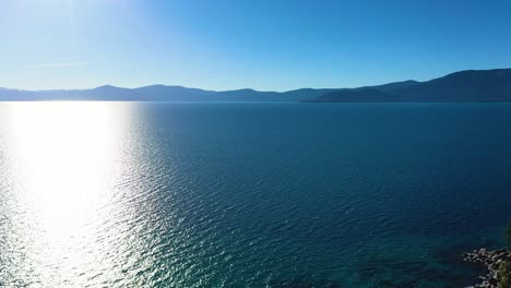 Wunderschöner-Lake-Taho-In-Kalifornien-Mit-Bergen-Im-Hintergrund-Und-Sonnenschein,-Der-Von-Der-Oberfläche-Glänzt,-Während-Der-Wind-Wellen-über-Blaues,-Kristallklares-Wasser-Mit-Einem-Felssteg-Am-Ufer-Kräuselt