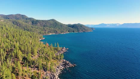 Aerial-Drone-Shot-of-Water-with-Crystal-Clear-Blue-Water-and-Thick-Pine-Tree-Forest-With-Mountain-Range-and-Rocky-Shoreline-at-Lake-Tahoe,-California