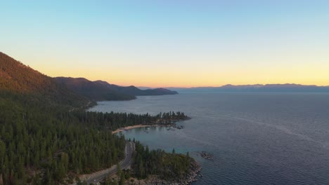 Colorful-Sunset-Over-Lake-Taho-with-a-Road-or-Highway-Going-Through-a-Pine-Tree-Forest-with-Clear-Blue-Water-and-Mountains-in-the-Background---aerial-drone-view