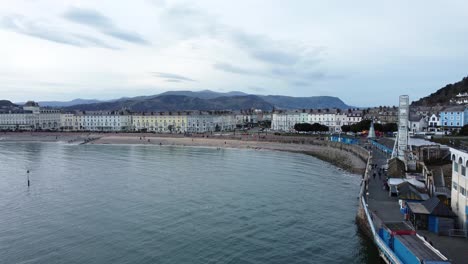 Aerial-view-Llandudno-Victorian-seafront-town-pier-promenade-passing-close-to-Grand-hotel-and-Ferris-wheel-boardwalk-attraction