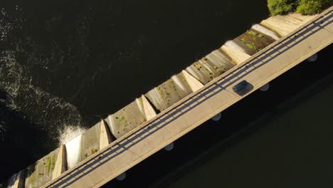 Aerial-top-down-shot-of-vehicle-driving-on-dam-road-crossing-lake-during-sunny-day-in-Argentina