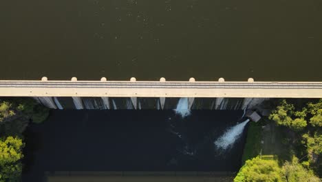 Aerial-top-down-shot-of-dam-construction-with-waterfall-crashing-into-river-and-tranquil-lake-in-sunlight