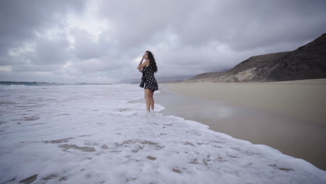 Multiracial-mixed-race-young-latin-woman-walking-alone-on-ocean-sand-beach-in-Fuerteventura-canary-island-Spain,-creative-photographer-exploring-new-touristic-spot