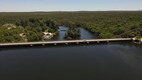 Aerial-view-showing-car-crossing-lake-by-dam-bridge-in-Cordoba,Argentina---Beautiful-forest-landscape-in-backdrop-during-summer