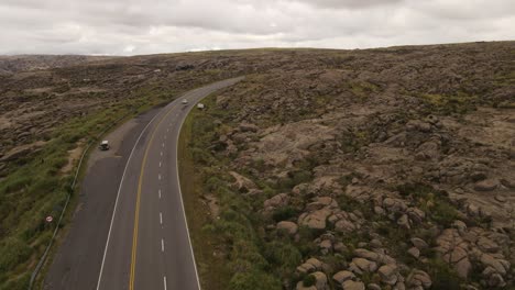Aerial-following-shot-of-white-car-driving-on-rural-road-surrounded-by-argentinian-highland-scenery-during-cloudy-day
