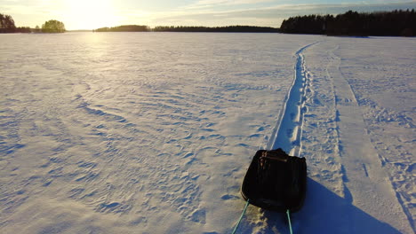 POV-backward-walking-and-dragging-sled-on-a-snowy-ice-covered-lake-by-sunset-golden-hour