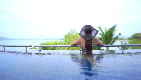 A-young-woman-with-her-back-to-the-camera-leans-along-the-swimming-pool-railing-as-she-looks-out-of-the-oceanview