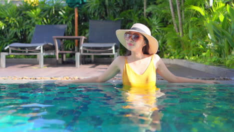 Hübsche-Asiatische-Frau-Mit-Sonnenbrille-Und-Strohhut,-Die-Im-Gelben-Badeanzug-Im-Pool-Steht,-Genießt-Die-Sonne-Im-Urlaub