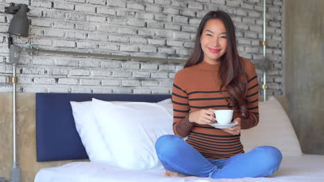 Close-up-of-a-young-woman-in-jeans-and-a-sweater-on-a-bed-holding-a-cup-of-coffee