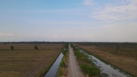 Aerial-reverse-footage-of-a-provincial-farm-road-revealing-canals-and-grassland,-Pak-Pli,-Nakhon-Nayok,-Thailand