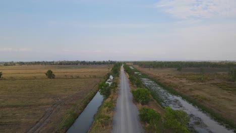 Descending-aerial-footage-revealing-this-farm-road-with-canals-for-irrigation,-Pak-Pli,-Nakhon-Nayok,-Thailand