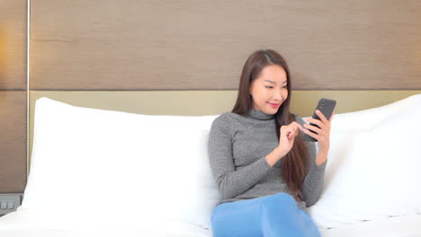 A-young-woman-sits-propped-up-in-a-bed-as-she-inputs-text-into-her-smartphone-then-celebrates-her-success