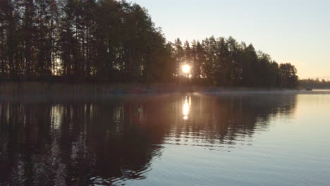 Rising-sun-flares-behind-trees-on-a-beautiful-misty-morning-in-calm-lake-scenery