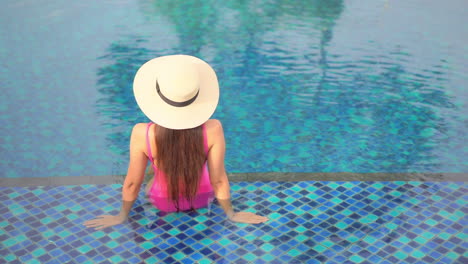 A-woman-with-her-back-to-the-camera-sits-on-the-shallow-step-of-a-swimming-pool