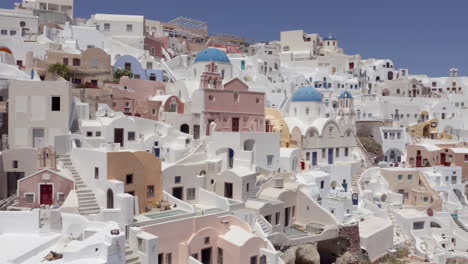 Aerial:-pullback-panoramic-shot-of-Oia-churches-in-Santorini,-Greece-on-a-sunny-day
