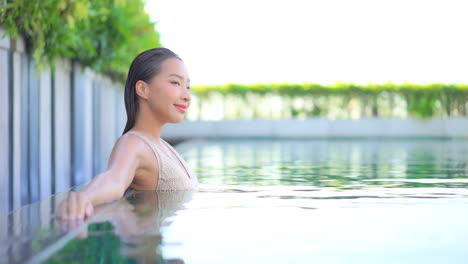 Happy-smiling-Asian-woman-standing-in-calm-swimming-pool-enjoying-vacation-and-resort-landscape