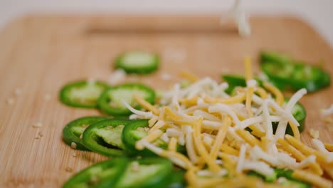 Multi-colored-shredded-cheese-dropping-on-bright-green-sliced-jalapeños-on-a-cutting-board-in-slow-motion