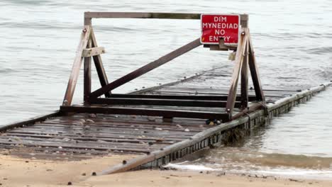 Tide-in-on-flooded-seaside-wooden-plank-walkway-jetty-with-Welsh-caution-warning-sign