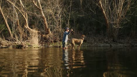 Männlicher-Fischer-Und-Sein-Hund-Stehen-Während-Der-Goldenen-Stunde-Mitten-Im-Fluss-Und-Werfen-Bei-Sonnenuntergang-Eine-Angelrute