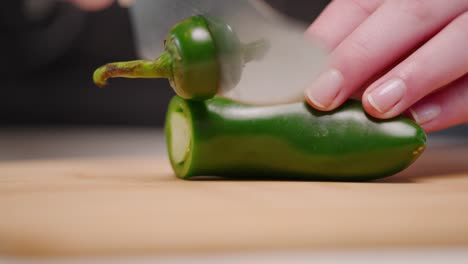 Caucasian-hand-cutting-whole-green-jalapeño-pepper-with-sharp-knife-onto-cutting-board-into-pile-of-slices-in-slow-motion