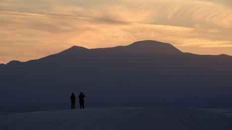Silhouetted-couple-on-sand-dune-watches-sunset-over-mountains-in-distance,-4K