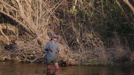 Male-fly-fisherman-in-hat-and-sunglasses-casts-his-fishing-rod-while-knee-deep-in-water-during-golden-hour-in-slow-motion