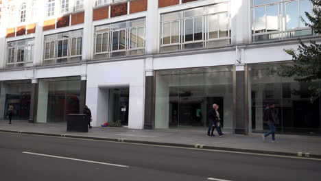 In-slow-motion-people-walk-past-a-homeless-person-sleeping-in-the-doorway-of-an-empty-retail-space,-known-as-“dead-spaces”,-on-Tottenham-Court-Road