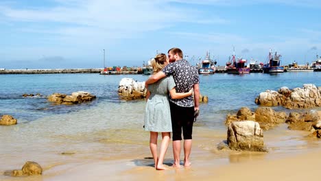 Romantic-young-couple-embrace-barefoot-in-warm-South-Africa-ocean-watching-Kalk-Bay-harbour-boats