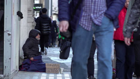 People-walk-past-a-homeless-person-sat-on-Oxford-Street-begging-for-money-with-a-cardboard-sign-that-reads,-“I-am-very-hungry,-God-bless”