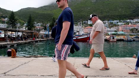 Casual-male-arriving-at-Kalk-bay-harbour-preparing-to-go-fishing-as-tourist-walk-through-scene