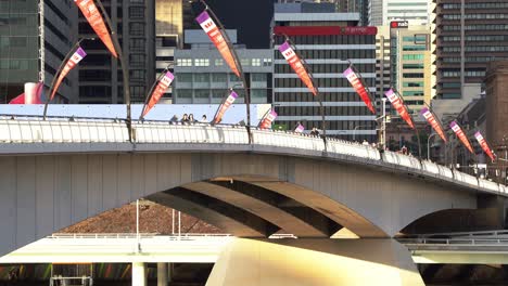 Pedestrians-stroll-across-Victoria-bridge-heading-to-South-Bank-at-sunset-golden-hours-with-buses-crossing-the-bridge-and-westpac-advertising-banners-waving-along-the-bridge,-establishing-static-shot