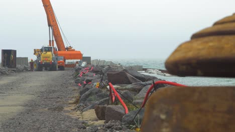 Construction-site-on-the-Baltic-sea-shore,-strengthening-the-Baltic-Sea-coastline,-building-protective-stone-pier,-heavy-duty-machinery-unloading-concrete-blocks,-overcast-day