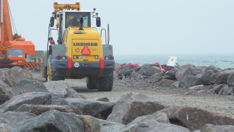 Construction-site-on-the-Baltic-sea-shore,-strengthening-the-Baltic-Sea-coastline,-building-protective-stone-pier,-yellow-heavy-duty-mover-driving-on-the-road,-overcast-day