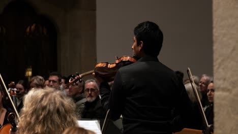 Medium-slow-motion-handheld-shot-of-an-orchestra-during-a-concert-with-violinists-playing-the-Mozart-Requiem-while-visitors-listen-to-the-sound
