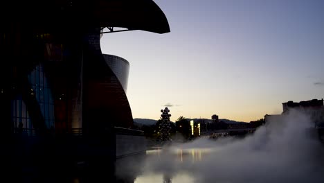 Guggenheim-Museum-Mit-Wassernebel-Und-Hohem-Baum-Und-Der-Augenskulptur-Von-Anish-Kapoor-Draußen-Bei-Sonnenuntergang,-Totale