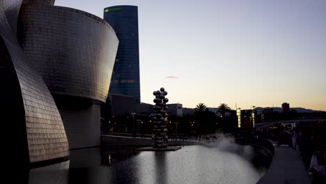 Metal-sheet-Guggenheim-Museum-with-water-mist-and-Tall-Tree-and-the-Eye-sculpture-by-Anish-Kapoor-outside-building,-Wide-shot