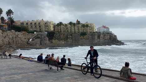 Azarujinha-Beach-with-a-very-rough-sea,-several-people-watch-the-waves-of-the-sea,-in-the-background-a-high-cliff-with-some-beautiful-housing-buildings