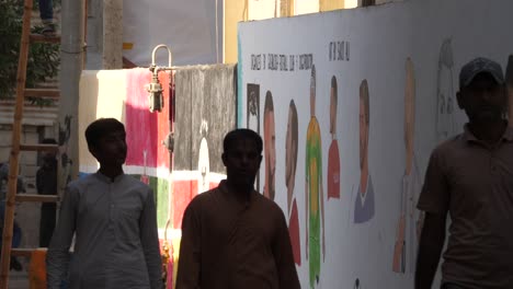 Locals-Walking-Past-Large-Wall-With-Paintings-Of-Football-Players-Celebrating-the-2022-World-Cup-In-Malir,-Karachi