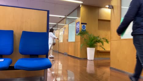 Older-doctor-walking-in-white-coat-and-mask-down-the-hallway-of-a-medical-clinic-from-the-waiting-room