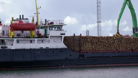 Crane-loads-timber-logs-onto-cargo-ship,-camera-pans-right,-mid-shot